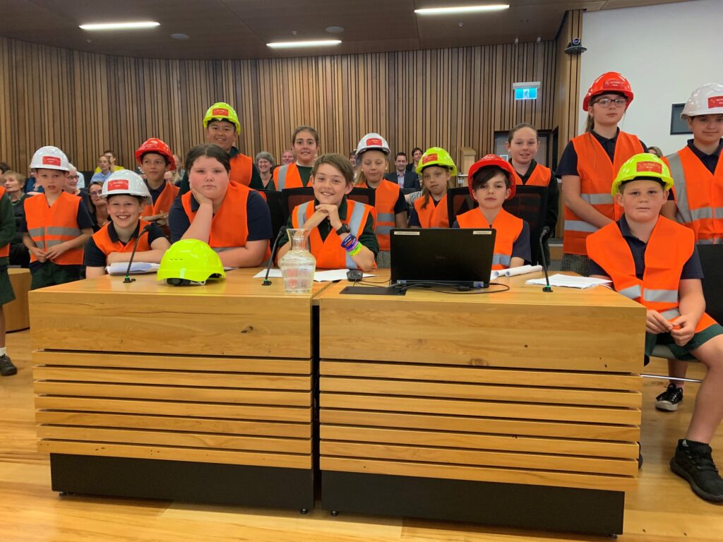 Group of children smiling at the council office behind a wooden desk, wearing hard hats and Hi-vis
